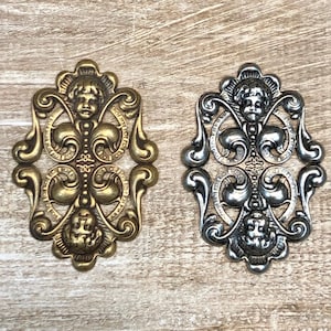 May include: Two ornate metal embellishments with a cherub design. One is gold and the other is silver.