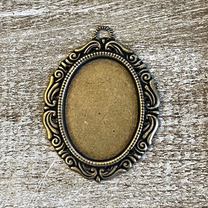 May include: A gold-toned metal pendant with an oval-shaped blank space in the center. The pendant has a decorative, ornate design around the edge.