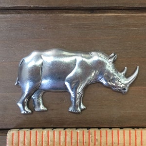 May include: A silver-colored rhinoceros figurine. The figurine is about 2 inches long and is standing on a wooden surface.