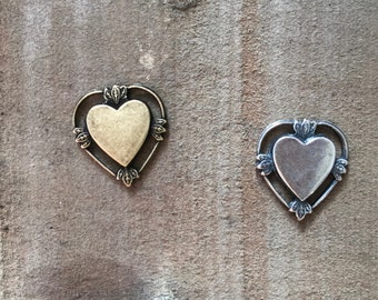 May include: Two heart-shaped metal pendants. One is gold-toned, the other silver-toned. Each has a heart-shaped center within a decorative frame with leaf accents. The pendants are displayed on a textured, neutral-toned surface.