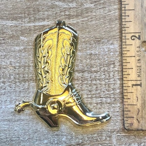 May include: A gold-colored cowboy boot ornament with a spur. The boot has a detailed design on the shaft and a buckle on the side.