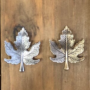May include: Two metal maple leaf charms, one silver and one gold, on a wooden surface.