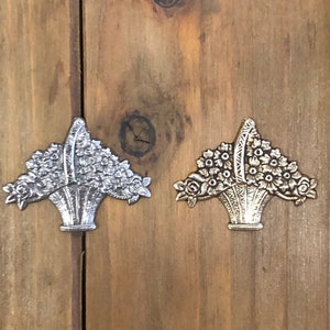 May include: Two silver metal flower baskets with a floral design. The baskets are on a wooden surface.