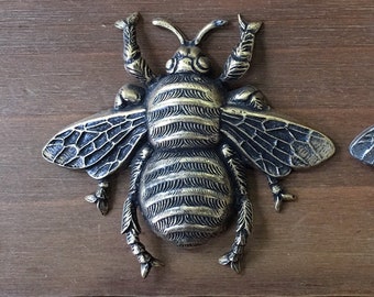 May include: Two metal bee figurines, one gold and one silver, are shown on a wooden surface. The bees are about 2 inches long and have detailed wings and legs.