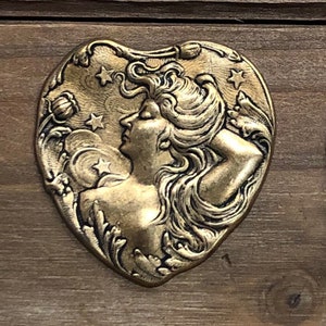 May include: Two heart-shaped metal charms featuring an Art Nouveau style woman with flowing hair and stars. The charms are approximately 1 inch in diameter and are shown next to a ruler.
