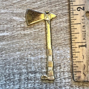 May include: A gold-colored miniature hatchet pin. The hatchet head and handle are detailed with a textured design. The pin is approximately 2 inches long, as measured by a wooden ruler in the image. The pin is set against a wood grain background.