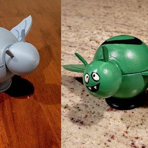 May include: A 3D-printed, light gray, spherical object with wing-like extensions and a smaller sphere attached. The object is displayed on a wooden surface. Next to it is a painted green version with a cartoon face.