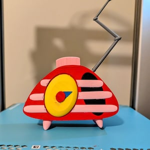 May include: A red, triangular-shaped radio figurine with a yellow circular dial, a blue triangular pointer, and a red center knob. It features horizontal light pink stripes, a black speaker area, three light pink legs, and a gray zigzag antenna.