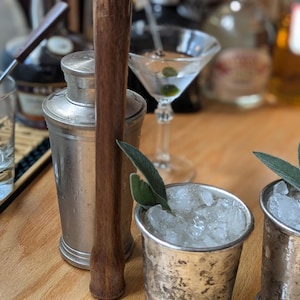 May include: Two silver metal cups filled with ice and a sprig of sage leaves. A wooden cocktail muddler and a silver cocktail shaker are also in the image.