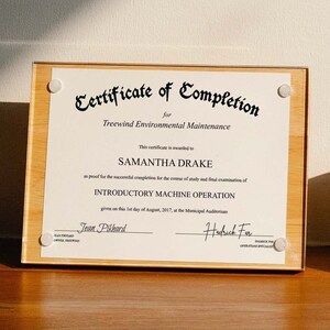 May include: A wooden certificate of completion for Treewind Environmental Maintenance is displayed on a wooden table. The certificate is awarded to Samantha Drake for completing the Introductory Machine Operation course. A white ceramic mug sits on a wooden coaster next to the certificate.