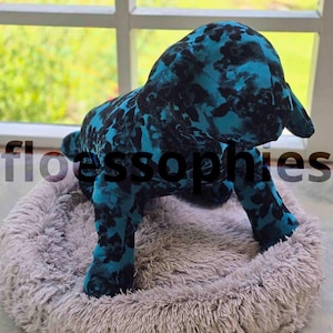 May include: A plush dog toy featuring a vibrant teal and black floral pattern, standing inside a round, shaggy, light beige pet bed. The word floessophies is printed in black across the centre of the image, with small text at the bottom right.