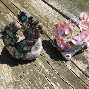 Op de afbeelding: Twee decoratieve voetsculpturen, één in iriserend groen en blauw, de andere in roze, zijn gemaakt van schelpen. Elk rust op een kleine, grijze steen, tegen een verweerd houten oppervlak.