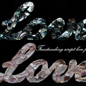 Puede incluir: Dos placas independientes con la palabra "LOVE" escrita en cursiva. La placa superior está hecha de piezas de concha iridiscentes azul-verdosas. La placa inferior está hecha de piezas de concha rosas y crema. El texto "Freestanding script love plaque" está escrito en cursiva.