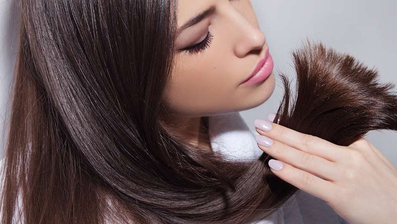 May include: A woman with long, dark brown hair is holding a section of her hair in her hand. She is looking down at her hair, and her expression is thoughtful. Her hair is very straight and shiny.