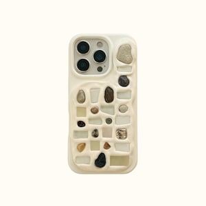 May include: A cream-colored smartphone case featuring a raised, grid-like texture embedded with various small, smooth, multi-colored stones and rectangular glass pieces. The design surrounds the camera module, creating a tactile, mosaic-like surface.