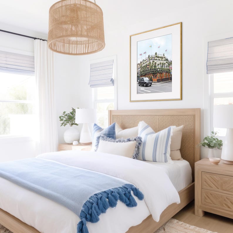 May include: A white bedroom with a bed with a blue blanket and white pillows. There is a framed print of a building with a car in front of it on the wall above the bed. There is a wicker lampshade hanging from the ceiling.