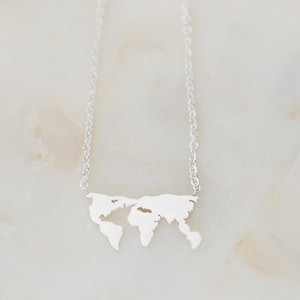 Puede incluir: Un collar plateado con un colgante de mapa del mundo. El colgante es un recorte detallado de los continentes, suspendido de una delicada cadena. El collar se muestra sobre una superficie clara.