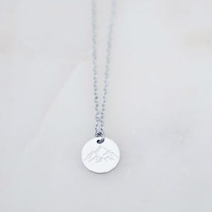 May include: Silver-toned necklace featuring a small, round pendant with a mountain range design. The pendant hangs from a delicate chain. The necklace is displayed against a white background.