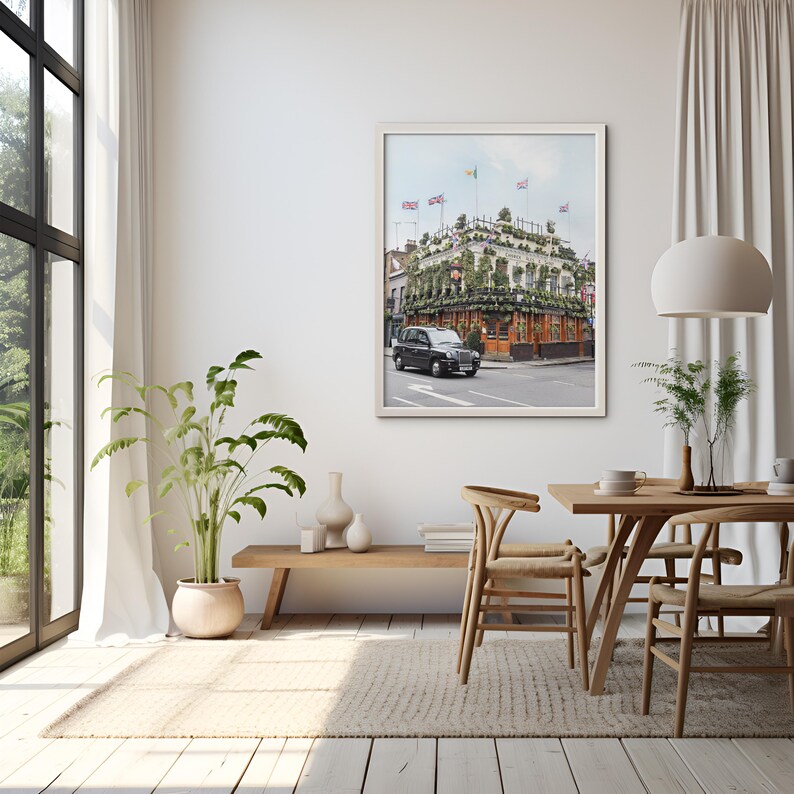 May include: A framed photograph of a black taxi cab parked in front of a building covered in greenery. The building has several British flags hanging from the roof. The photo is hanging on a white wall in a modern living room.