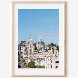 Puede incluir: Una impresión enmarcada de un paisaje urbano con una iglesia alta y ornamentada con agujas gemelas, sobre un cielo azul claro. La imagen captura una vista de una calle de la ciudad bordeada de edificios, con una mezcla de estilos arquitectónicos.