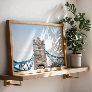 May include: A framed photograph of Tower Bridge in London, England. The photo is in a light wood frame and is hanging on a wooden shelf with gold metal brackets.