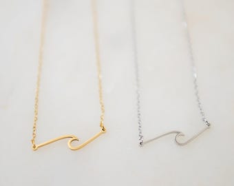 May include: Two delicate necklaces featuring a minimalist wave pendant. One necklace is finished in a bright gold tone, while the other is finished in a silver tone. Both necklaces consist of a thin, fine link chain attached to the wave charm.