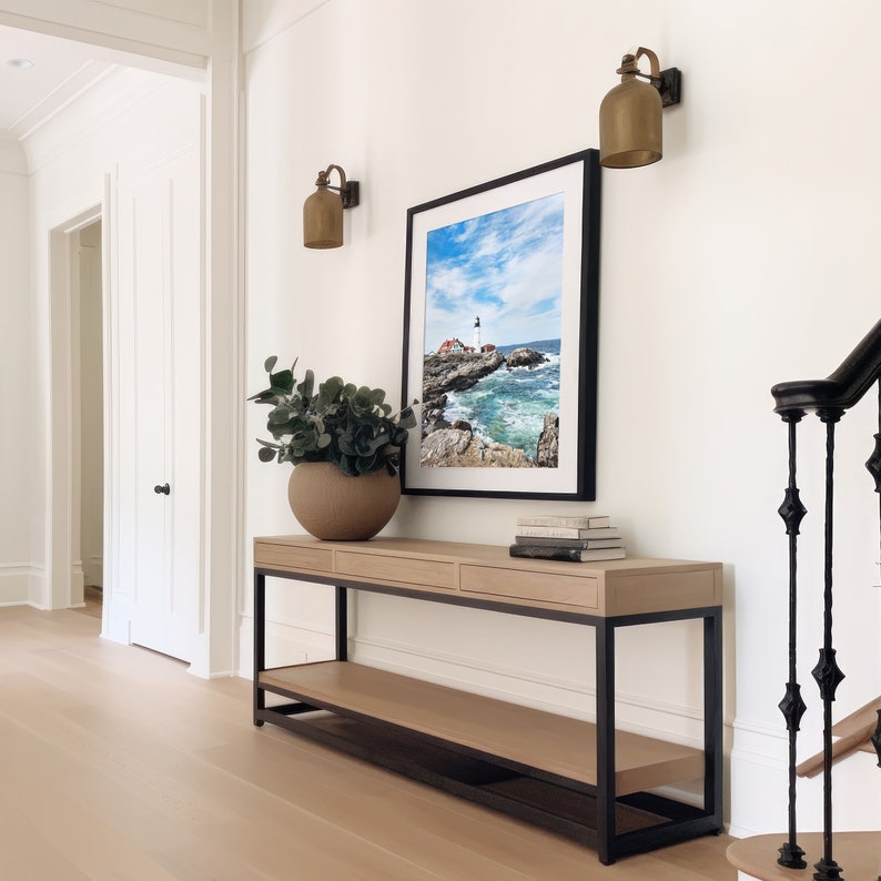 May include: A light wood console table with black metal legs and a lower shelf. The table has two drawers and is decorated with a potted plant, a framed photograph of a lighthouse, and a stack of books.