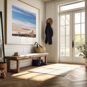 Pu&ograve; includere: Una panca di legno con un cuscino bianco si trova in un corridoio con una grande fotografia di paesaggio incorniciata sul muro, un cappello di paglia e una giacca blu navy appesi a un gancio, una borsa di paglia sulla panca e un tappeto bianco sul pavimento.
