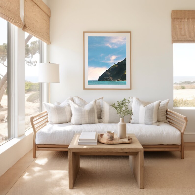 May include: A white couch with a wooden frame and light brown cushions sits in a living room with large windows. A framed photograph of a cliffside beach scene hangs on the wall above the couch. A small wooden coffee table sits in front of the couch with a tray of decorative items on top.