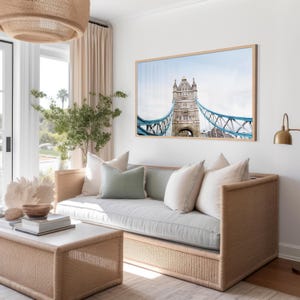 May include: A light gray sofa with a woven rattan frame and light blue cushions sits in a living room with a large framed print of the Tower Bridge in London above it.