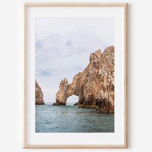 May include: A framed print showing a natural rock arch over the sea. The rock is a light brown colour, and the water is a deep blue-green. The sky is overcast with a light grey colour.