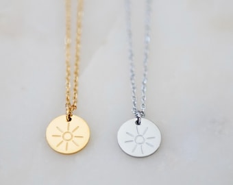 Puede incluir: Dos collares con cadenas metálicas finas y colgantes circulares. Un colgante es de tono dorado y el otro de tono plateado. Cada dije circular presenta un diseño de sol grabado con un círculo central y líneas radiantes.