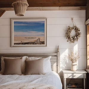 Pu&ograve; includere: Una camera da letto bianca con un letto, un comodino e una fotografia incorniciata di un paesaggio desertico. Il letto ha un piumone bianco e due cuscini marroni. Il comodino ha un vaso con rami secchi e un libro sopra. La fotografia &egrave; appesa al muro sopra il letto.