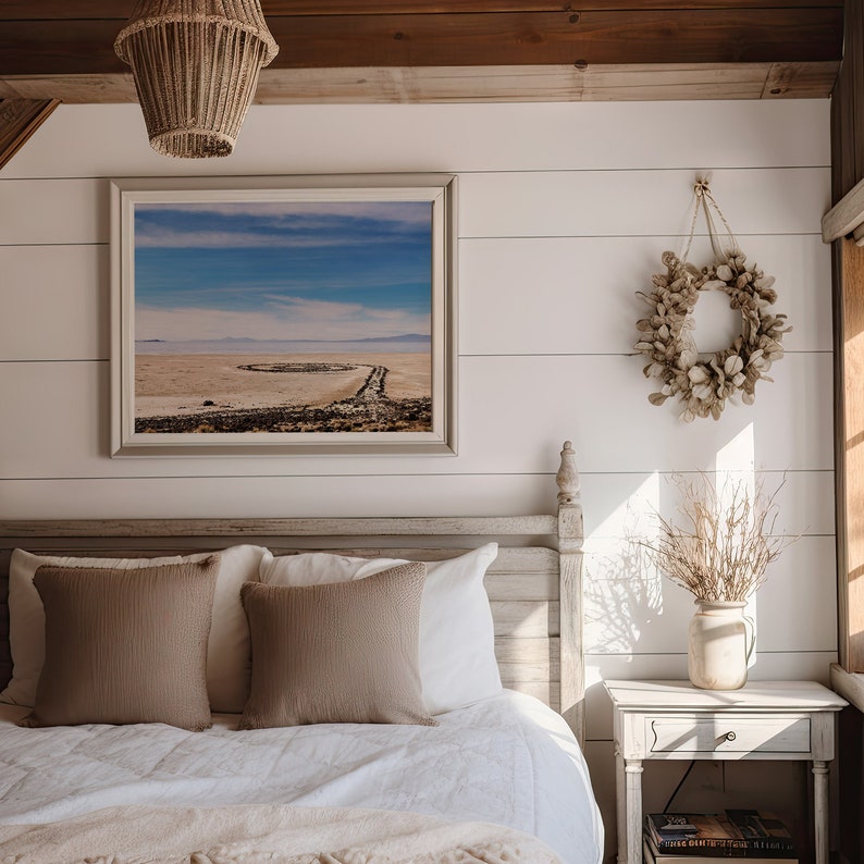 Pu&ograve; includere: Una camera da letto bianca con un letto, un comodino e una fotografia incorniciata di un paesaggio desertico. Il letto ha un piumone bianco e due cuscini marroni. Il comodino ha un vaso con rami secchi e un libro sopra. La fotografia &egrave; appesa al muro sopra il letto.