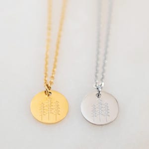 May include: Two necklaces with a simple design of three pine trees on a round pendant. One necklace has a gold chain and pendant, the other has a silver chain and pendant.