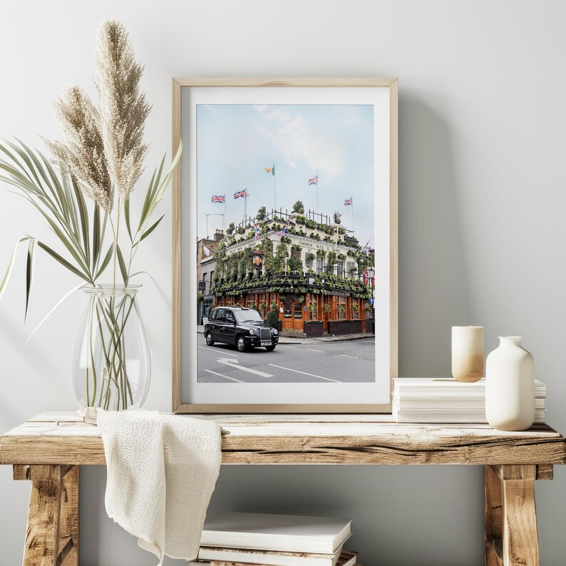 May include: A black London taxi cab parked in front of a building covered in greenery. The building has a red brick facade and is decorated with flags. The building is located on a street with other buildings in the background.