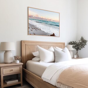 May include: A bedroom with a wooden bed frame and white bedding. A framed photograph of a beach scene hangs on the wall above the bed. There are two bedside tables with lamps and a plant on one of them.