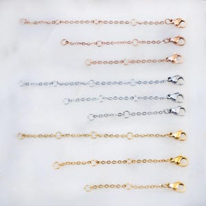 May include: An assortment of jewelry chain extenders in rose gold, silver, and gold tones. Each extender features a small chain with a lobster clasp and a jump ring for easy attachment to necklaces or bracelets.