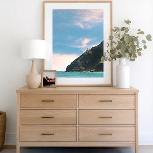 May include: A light wood dresser with six drawers and brass pulls. A framed photograph of a rocky coastline with a lighthouse on top of the cliff is hanging above the dresser. A white lamp with a white shade is on the left side of the dresser. A vase with green leaves is on the right side of the dresser.