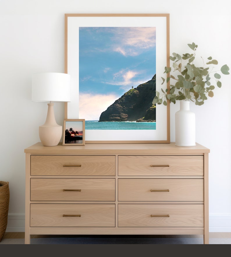 May include: A light wood dresser with six drawers and brass pulls. A framed photograph of a rocky coastline with a lighthouse on top of the cliff is hanging above the dresser. A white lamp with a white shade is on the left side of the dresser. A vase with green leaves is on the right side of the dresser.