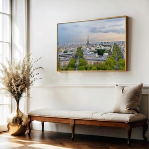 May include: A framed print of Paris, showing the Eiffel Tower and avenues lined with trees. The artwork has a gold frame and is displayed on a white wall above a wooden bench with a beige cushion. A gold vase with dried flowers is on the left.