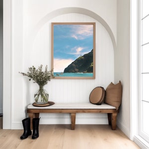 May include: A wooden bench with a white cushion and two black rain boots sits in a white room with a framed photograph of a rocky coastline and ocean.