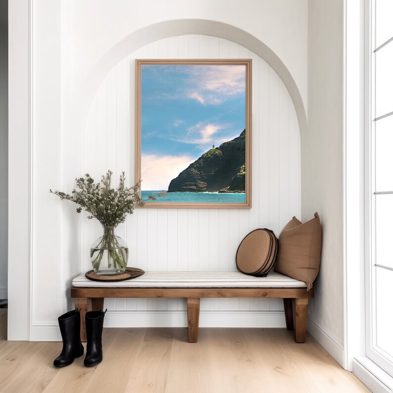 May include: A wooden bench with a white cushion and two black rain boots sits in a white room with a framed photograph of a rocky coastline and ocean.