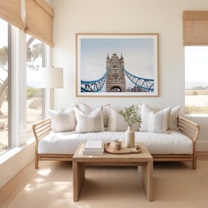 May include: A white couch with pillows sits in front of a window with a view of trees and a body of water. A framed photograph of the Tower Bridge in London hangs above the couch. A wooden coffee table sits in front of the couch with a tray, a cup, and a stack of books on top.