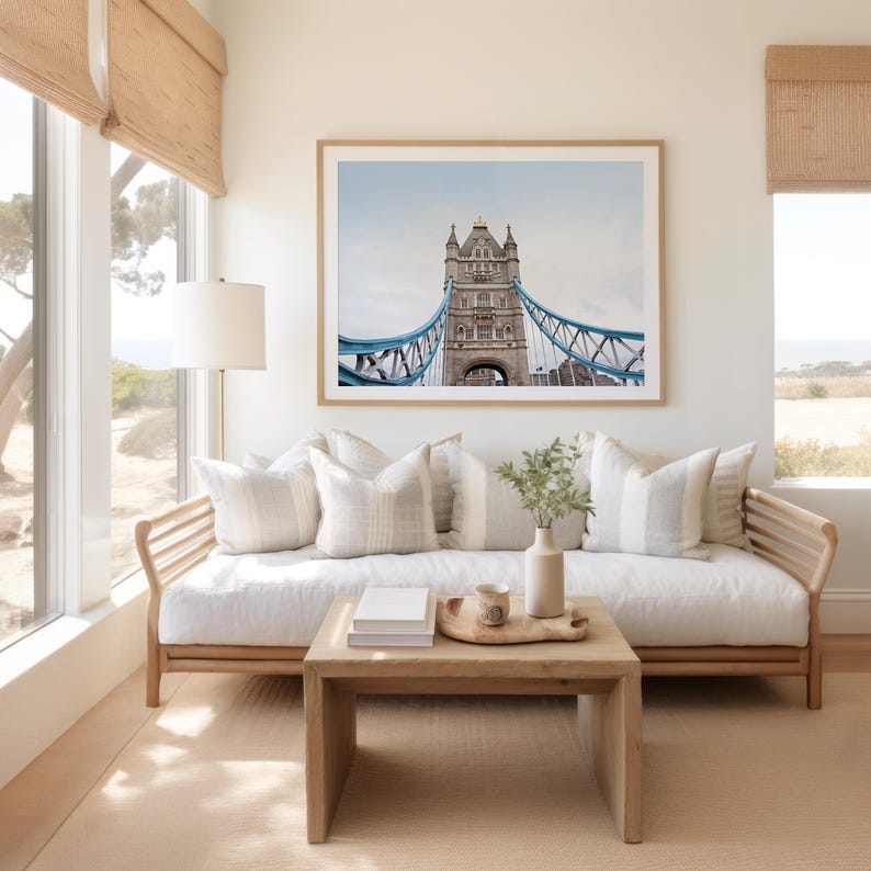 May include: A white couch with pillows sits in front of a window with a view of trees and a body of water. A framed photograph of the Tower Bridge in London hangs above the couch. A wooden coffee table sits in front of the couch with a tray, a cup, and a stack of books on top.