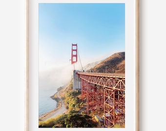 May include: Framed print of the Golden Gate Bridge in San Francisco. The iconic red bridge spans across the water, with a clear blue sky above. The bridge's towers and roadway are visible, along with the surrounding hills and coastline.