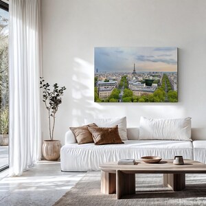 May include: A framed canvas print depicting Paris, France, with the Eiffel Tower and avenues lined with trees. The artwork is displayed above a white sofa with brown cushions. A wooden coffee table and a potted plant are in the foreground.