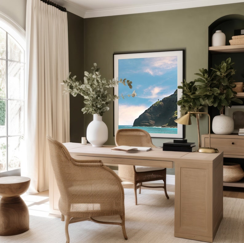 May include: A home office with a wooden desk, two woven chairs, and a large framed photograph of a tropical island landscape. The desk has a lamp, a vase with greenery, and a book on top. The walls are painted a muted green color.
