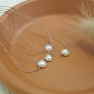 Puede incluir: Tres collares delicados, cada uno con una pequeña perla de agua dulce blanca de forma irregular suspendida en un fino cordón metálico trenzado. Los collares están dispuestos en capas, mostrando la textura brillante de las perlas.