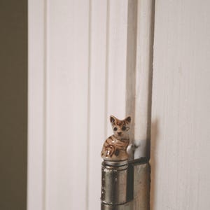 May include: A small, ceramic cat figurine with brown stripes and markings sits atop a metal hinge. The cat is detailed with a sitting pose. The background is a white door with vertical paneling.